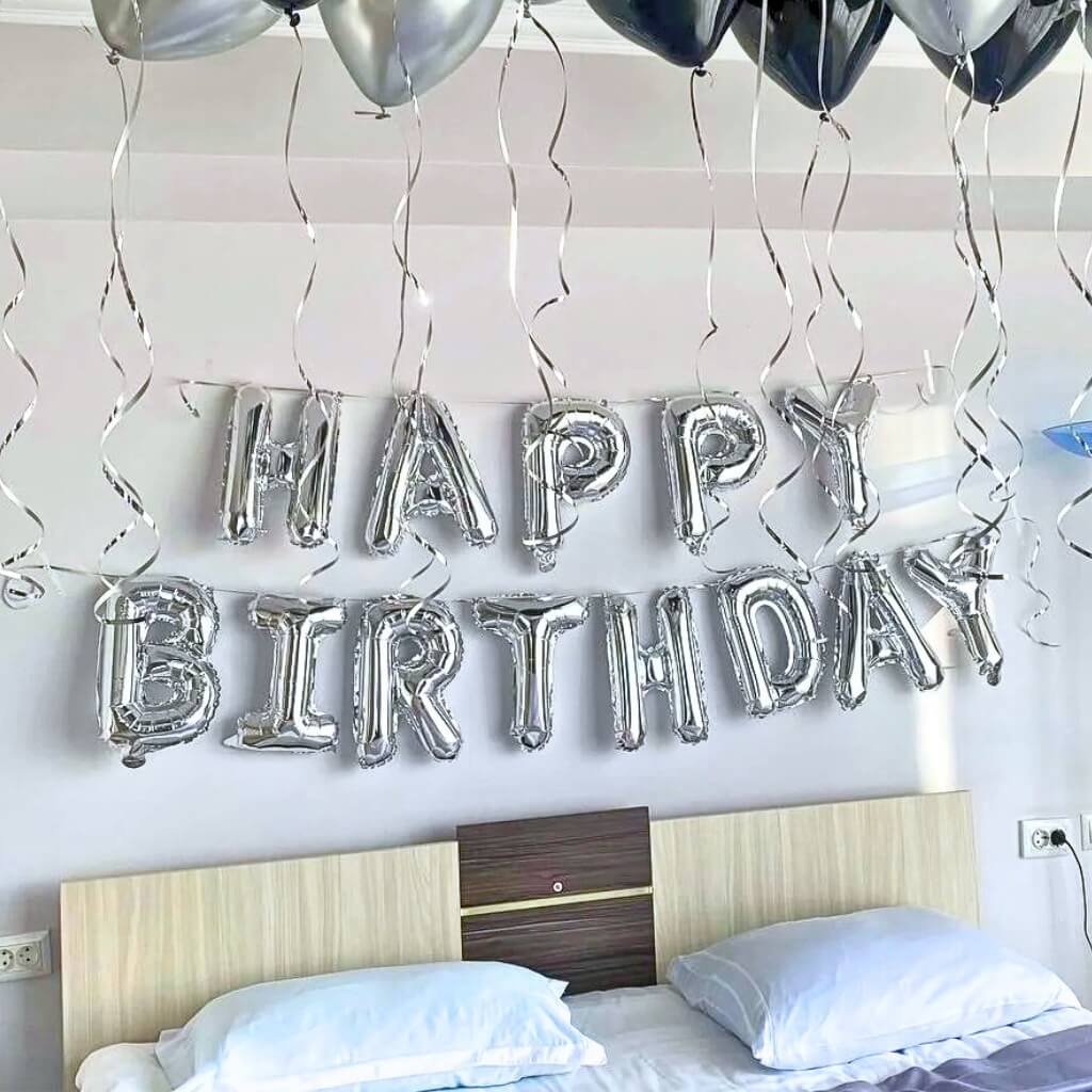 Silver 'Happy Birthday' balloon letters hanging above a bed with a neutral background
