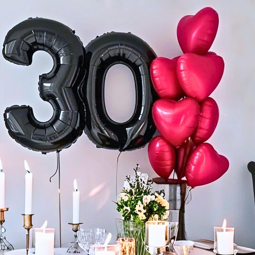 Black '30' foil balloons and matte red heart-shaped balloons on a table with candles and flowers.