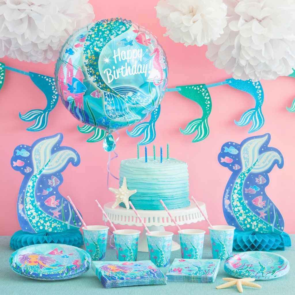 Mermaid-themed birthday party setup with cake, balloons, and decorations on a pink background.